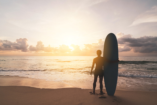 Man hold surfboard standing at tropical sunset beach background. Summer vacation and sport adventrue concept. Vintage tone filter effect color style.