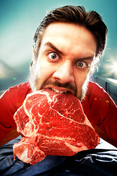 A man holding a large raw piece of meat with his teeth stock photo