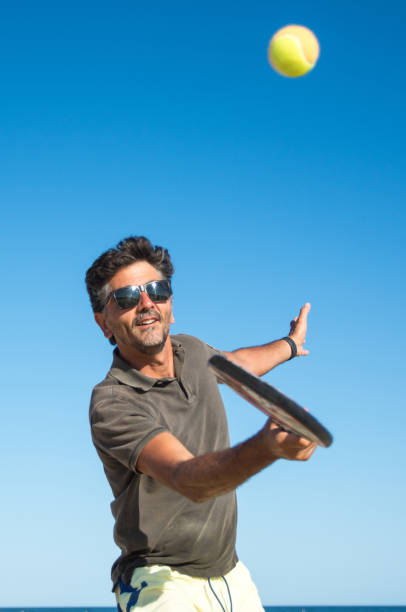 homem segurando uma raquete de tênis de remo batendo a bola em um fundo azul. - beach tennis - fotografias e filmes do acervo