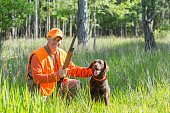 Hunter kneeling on the ground in tall grass, smiling at the camera holding a shotgun.  He is petting his hunting dog, a brown labrador retriever, sitting next to him.  He is wearing an orange safety vest.