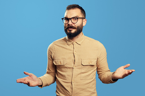 Young bearded man in bewilderment shrugging his shoulders and looking at camera with confusion, isolated over blue background