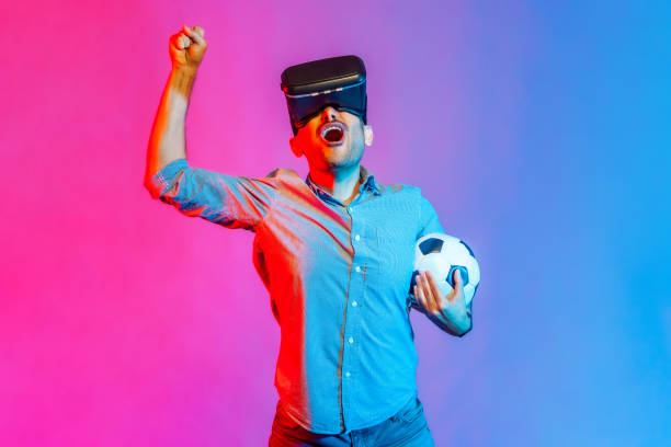 Man in headset and holding soccer ball, watching 3d football team, cheering, celebrating victory. stock photo