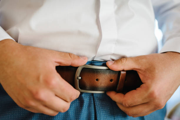 A man in pants and shirt buttoned a brown leather trouser belt. Groom dress up a belt with buckle. Businessman wear leather stylish belt. stock photo