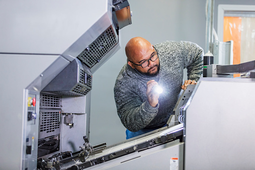 A mid adult man in his 30s working in a printing factory. He is looking into one of the printing presses with a flashlight, trying to fix a problem. He is mixed race African-American and Filipino.