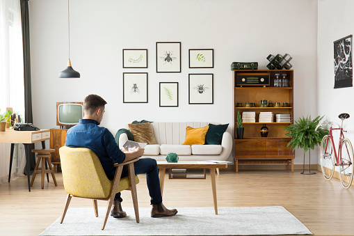 Man sitting on yellow chair at wooden table in retro living room with sideboard and gallery above settee