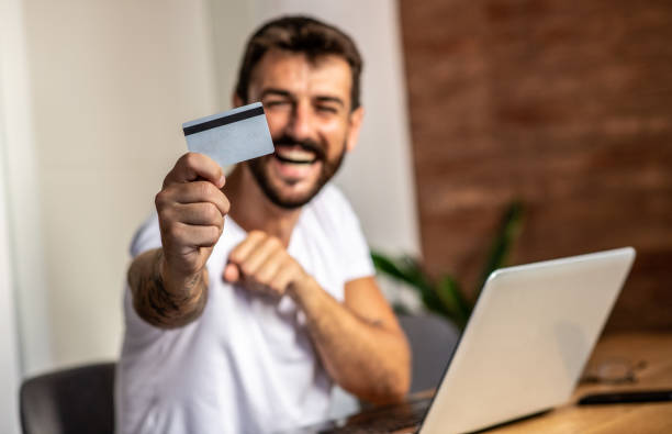 A man in the morning with a bank credit card in his hand, happy about a new online purchase, sitting at a table in the dining room stock photo