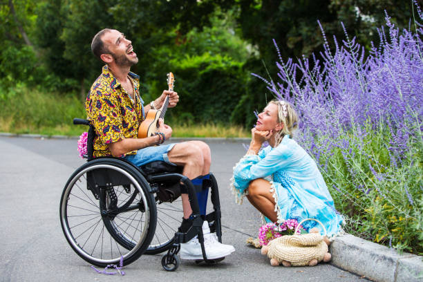 man in wheelchair outdoors playing the guitar for blond woman sitting at a curb - wheelchair street imagens e fotografias de stock