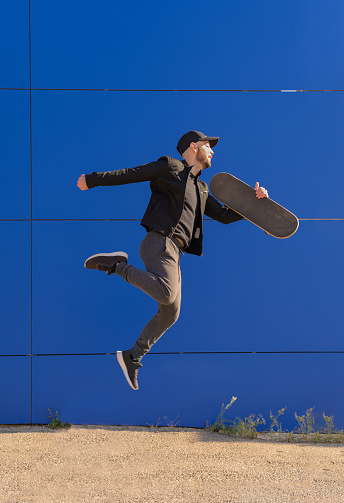 Full body of a young man in casual attire jumping high while performing a trick on a sunny day in the city with a blue wall in the background.
