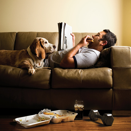 Hispanic man working out with weights laying down on a sofa reading a magazine with his dog.