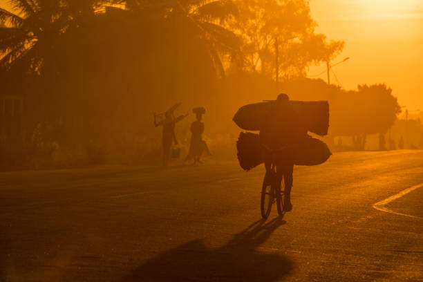 Man on a bicycle with bags at sunrise A Mozambican cyclist carrying large bags of coal to market at sunrise in the morning, along a tarmac road. Other commuters carrying items in the background. Nampula Town, Mozambique, Africa developing countries stock pictures, royalty-free photos & images