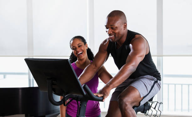 Man on exercise bike at home, supportive partner An African-American couple in their 30s exercising at home in the living room. The man is on an exercise bike and his partner is standing beside him, being supportive and encouraging. peloton stock pictures, royalty-free photos & images