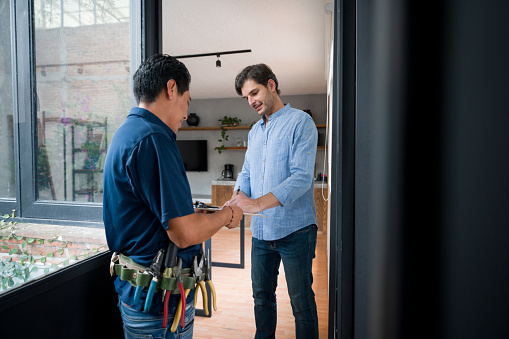 Latin American Man opening the door of his house to an electrician and signing a report after requesting a service - home improvement concepts