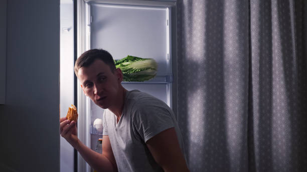 Man opens fridge door eats eclair in secret in dark kitchen stock photo