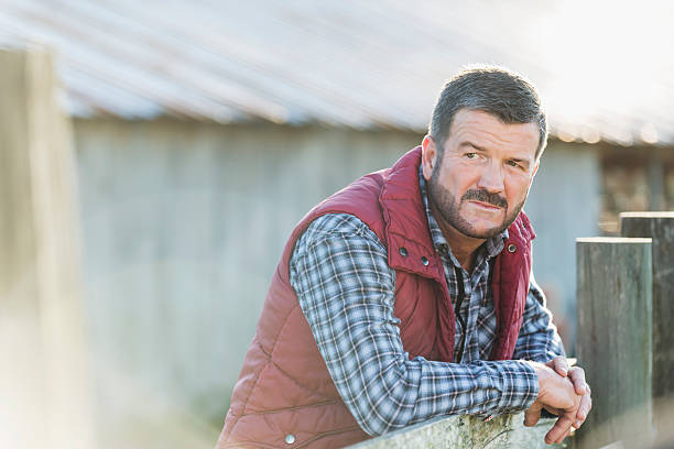 Man outside barn leaning on wooden fence A farmer or rancher, mature man in his 50s, standing outside a barn, with his arms leaning on a wooden fence. He is wearing a plaid shirt and warm vest. rancher stock pictures, royalty-free photos & images