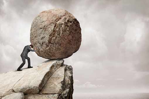 A businessman attempts to push a giant round boulder off of a cliff.  The monochrome background and ky provide ample negative space for copy and text.