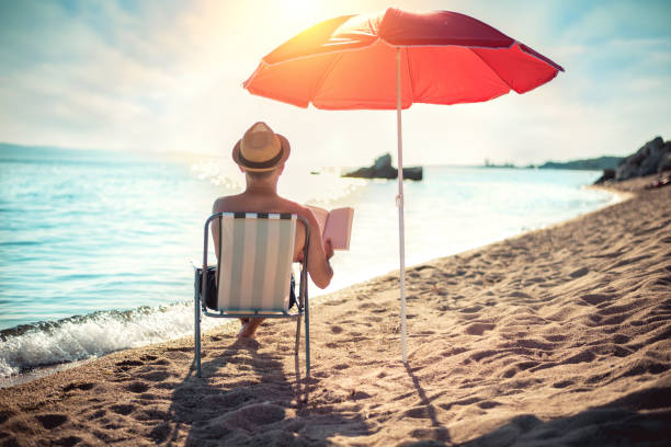 man läser bok i solstol under solparasoll nära havet - parasol bildbanksfoton och bilder