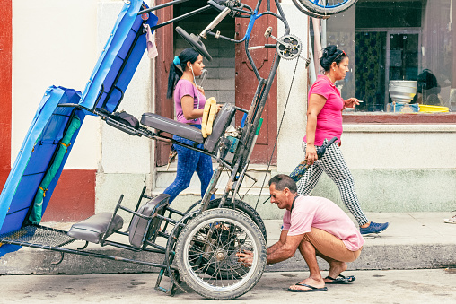 Santa Clara, Villa Clara, Cuba - November 8, 2019: A Cuban man repairs a bicitaxi or pedicab by the sidewalk where two women are seen walking. Bicitaxis are privately operated small businesses for urban transportation.