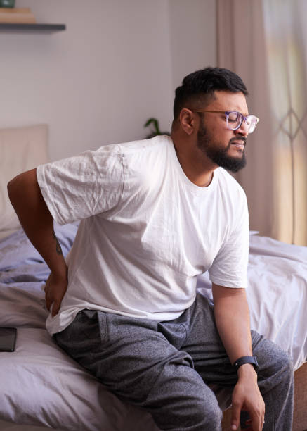 A man sits on the edge of the bed holding his lower back in pain stock photo