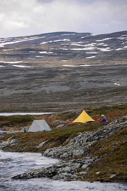 Man sitting by tents and river in wilderness with mountains A male backpacker rests by a river in the Lapland wilderness. Yellow and green tents are pitched nearby. kilpisjarvi lake stock pictures, royalty-free photos & images