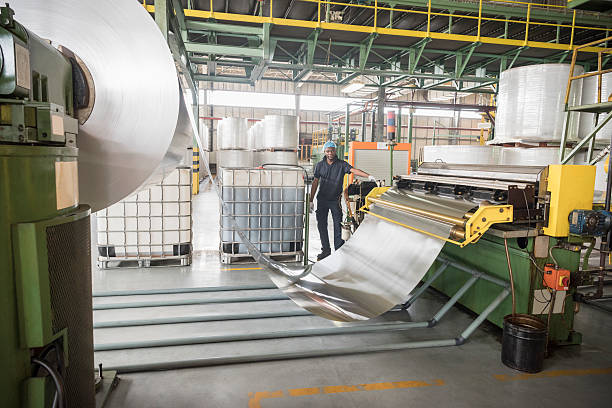 Man standing by large roll of aluminium in factory Male factory worker in aluminium processing plant. Rolls of aluminium metal being processed inside industrial building. sheet metal stock pictures, royalty-free photos & images