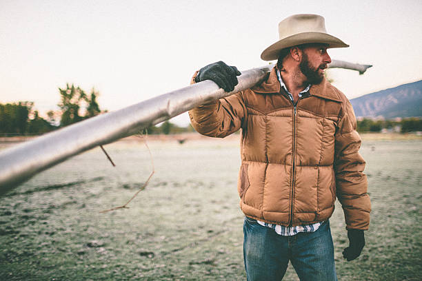 Man standing in field carries irrigation pipe over shoulder Man standing in field carries irrigation pipe over shoulder rancher stock pictures, royalty-free photos & images