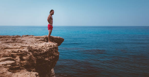 Man standing on cliff edge ready to jump into sea Young active diver standing on cliff looking at sea and preparing to dive into ocean cliff jumping stock pictures, royalty-free photos & images