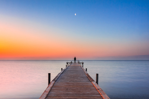 Man standing on jetty at beach with sunrise and moon