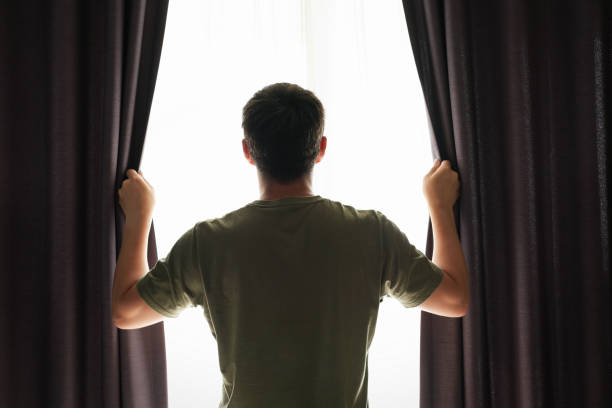 Man stands at the window and opens curtains back view. stock photo