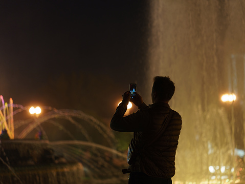 Moscow, Russia - July 21, 2019, Long exposure image of the sprays of colorful fountain in night time. Silhouette of young man taking photography of it.