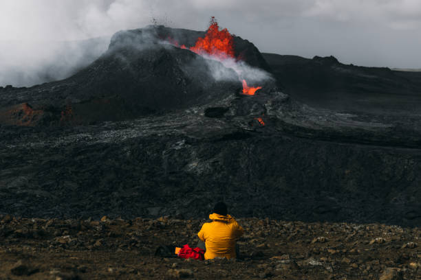 Man traveler feeling awe looking at Fagradalsfjall volcanic eruption in Iceland Young man backpacker in yellow jacket exploring the dramatic landscape, sitting near the big erupting volcano with melting lava in Iceland active volcano stock pictures, royalty-free photos & images