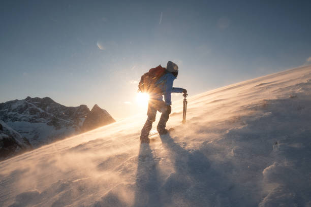 Photo of Man traveler with tripod are climbing on slope hill on Ryten mount in blizzard at sunset