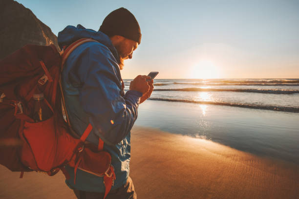 Man traveling with backpack using smartphone on beach navigation application modern technology influencer lifestyle active vacations outdoor sunset sea view stock photo