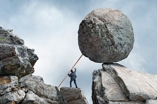 A businessman uses a long piece of lumber in an attempt to support a large round boulder and keep it from crashing down on him.