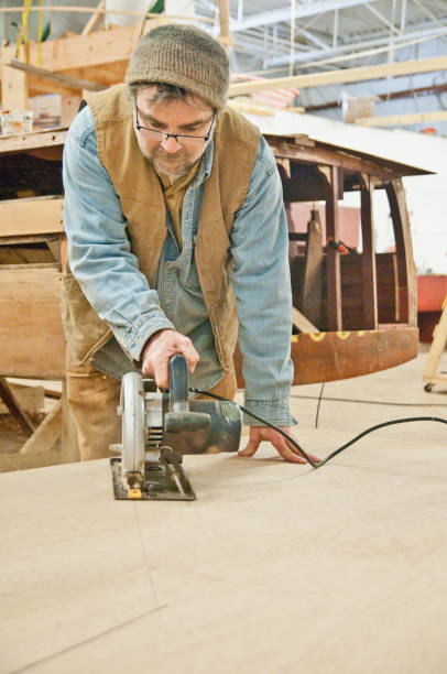 Man uses circular saw on plywood in wooden boat shop stock photo
