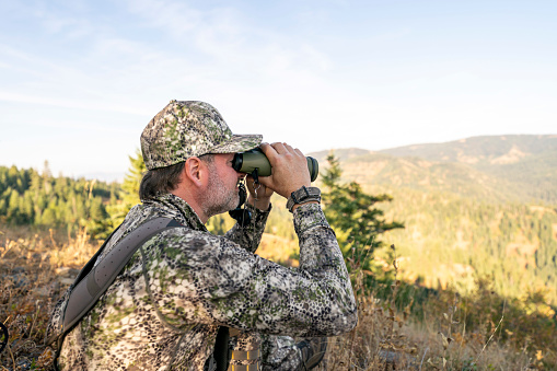 A hunter sits on a mountain peak and looks through binoculars while tracking wild game in the forested wilderness of Washington State.