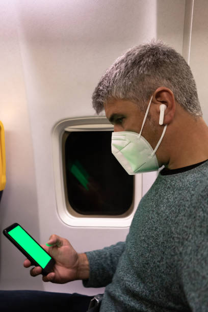 Man using smart phone with green screen wearing a face mask and earphones in airplane seat stock photo