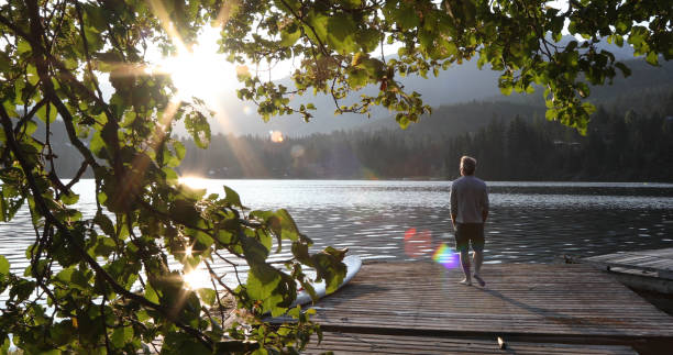 Man walks onto dock over lake and watches sunrise over mountains and forest He looks off to distant scene barefoot photos stock pictures, royalty-free photos & images