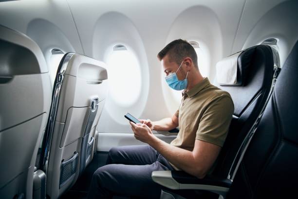 Man wearing face mask inside airplane Man wearing face mask and using phone inside airplane during flight. Themes new normal, coronavirus and personal protection. passenger stock pictures, royalty-free photos & images