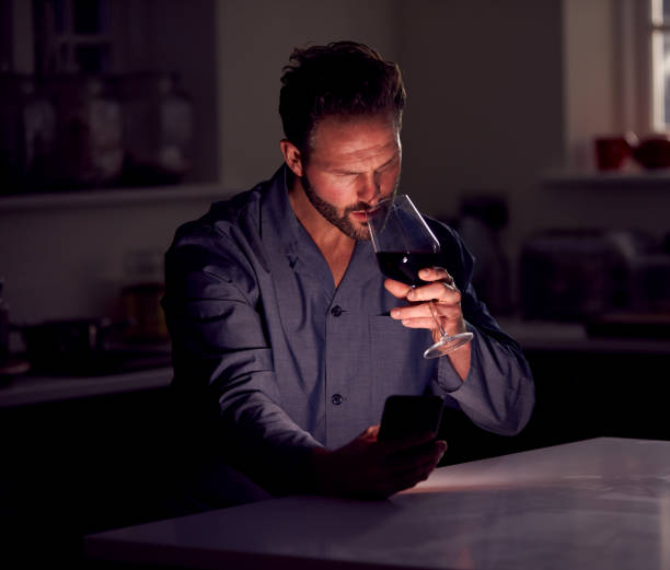 Man Wearing Pyjamas Sitting In Kitchen With Glass Of Wine At Night Using Mobile Phone Man Wearing Pyjamas Sitting In Kitchen With Glass Of Wine At Night Using Mobile Phone fomo photos stock pictures, royalty-free photos & images