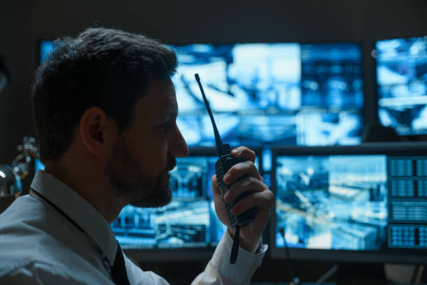 A man with a beard, a security guard or a police officer, possibly from the rescue service, is talking on a walkie-talkie against the background of digital monitors that display information from surveillance cameras. stock photo