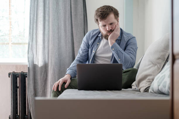 Man with a laptop is bored while sitting at home. He is sad because he is forced to stay home during a virus and pandemic epidemic. stock photo