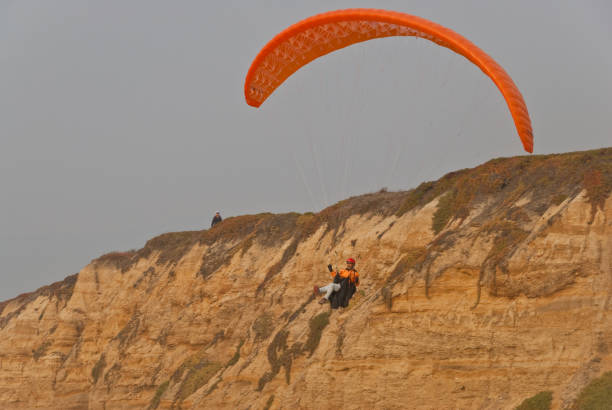 Paragliding Off a Bluff Half Moon Bay, California, USA - November 09, 2018: A man with an orange paraglider jumps off a bluff to land on the Pacific beach. jeff goulden paragliding stock pictures, royalty-free photos & images