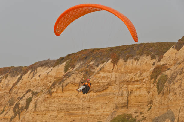 Paragliding Off a Bluff Half Moon Bay, California, USA - November 09, 2018: A man with an orange paraglider jumps off a bluff to land on the Pacific beach. jeff goulden paragliding stock pictures, royalty-free photos & images