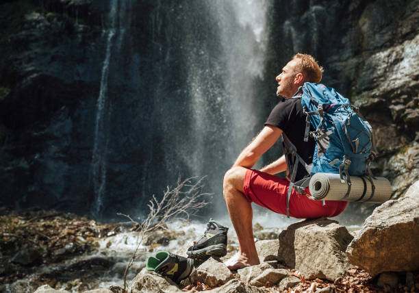 Man with backpack dressed in active trekking clothes takes off trekking boots sitting near mountain river waterfall and enjoying the Nature. Traveling, trekking, or nature concept image stock photo