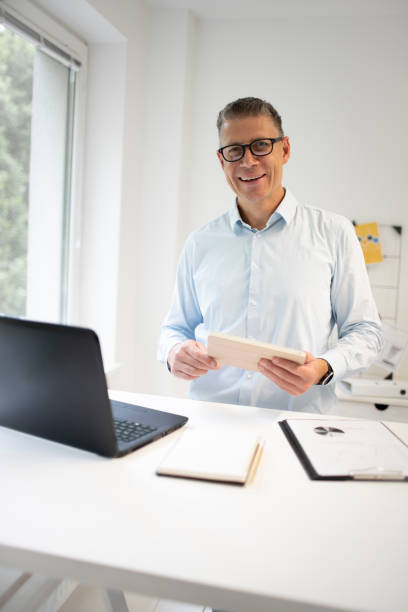 man with blue shirt and black glasses is standing behind standing table and is working with his tablet stock photo