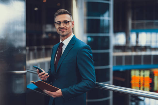 Man with documents and cellular looking at camera and smiling Attractive, adult man in chic formal wear standing inside business center. He looking at camera and holding telephone with document in hands businessman stock pictures, royalty-free photos & images