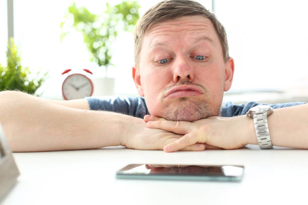 Man with funny facial expression staring at cellphone Man with funny facial expression staring at cellphone lying at table waiting for call concept waiting stock pictures, royalty-free photos & images