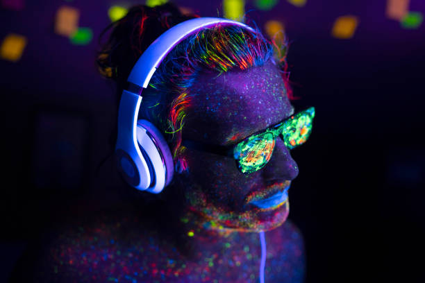 Man with headset and sunglasses painted in fluorescent UV colors Man with glowing makeup in black light. Man with neon makeup powder on face. Man painted in fluorescent UV colors, with sunglasses and headset. paint neon color neon light ultraviolet light stock pictures, royalty-free photos & images