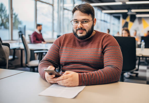 Man working at  the office One young overweight man with reading glasses sitting at the office, using smart phone and reading documents. fat man looks at the phone stock pictures, royalty-free photos & images