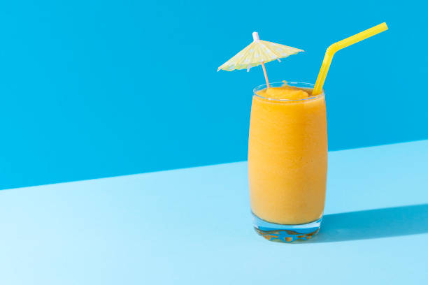 Mango smoothie with a cocktail umbrella. Summer cold drink Glass of mango smoothie with summer decor on a blue background, in harsh light. Healthy smoothie drink. Vitamin breakfast beverage. Dietary mocktail. mango smoothie stock pictures, royalty-free photos & images